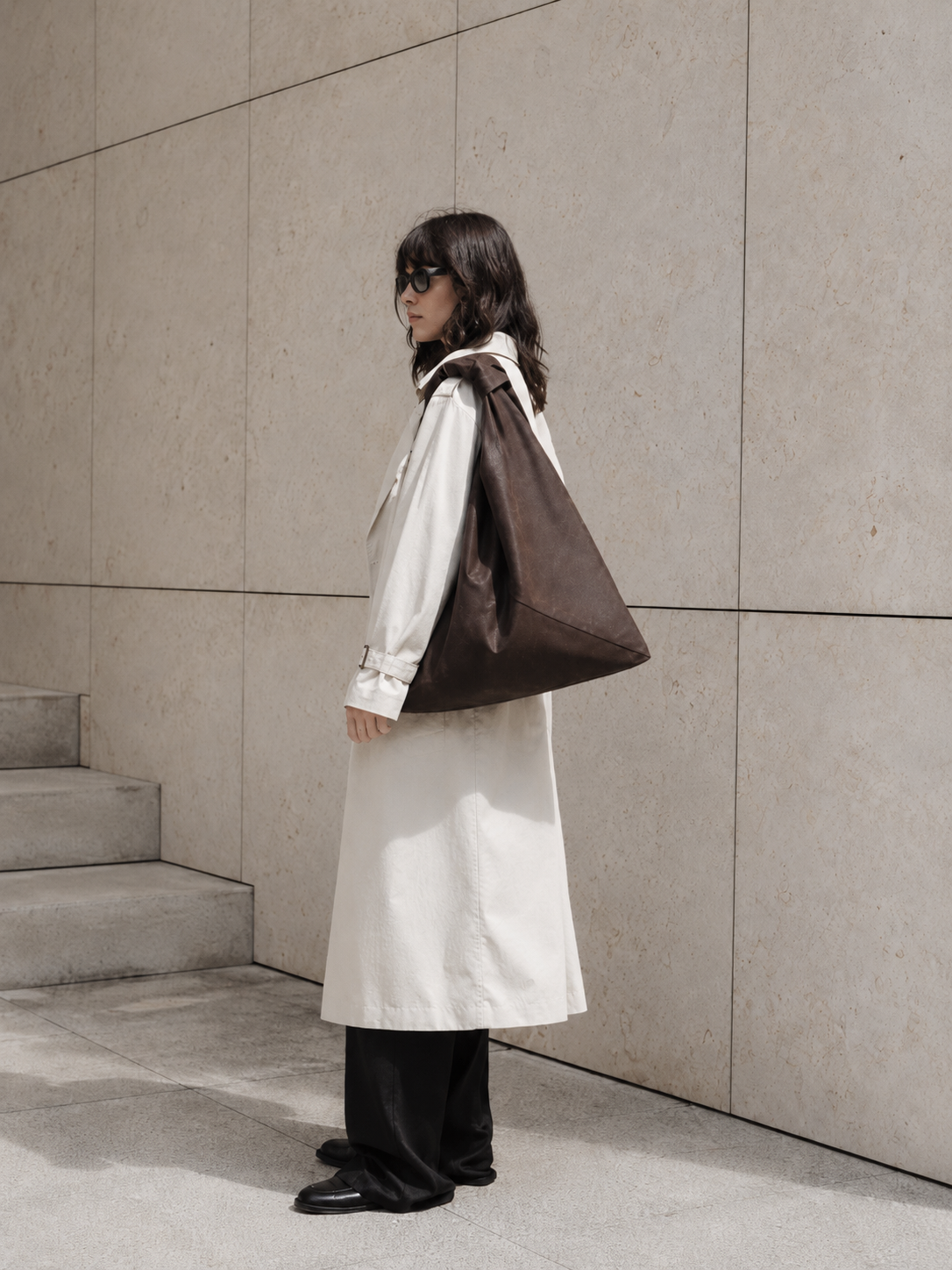 A woman stands in profile against a beige stone wall, wearing a long cream coat over a white top, black wide-leg pants, and black shoes, while carrying a large dark brown waxed canvas origami bag made by Esra Kayira.