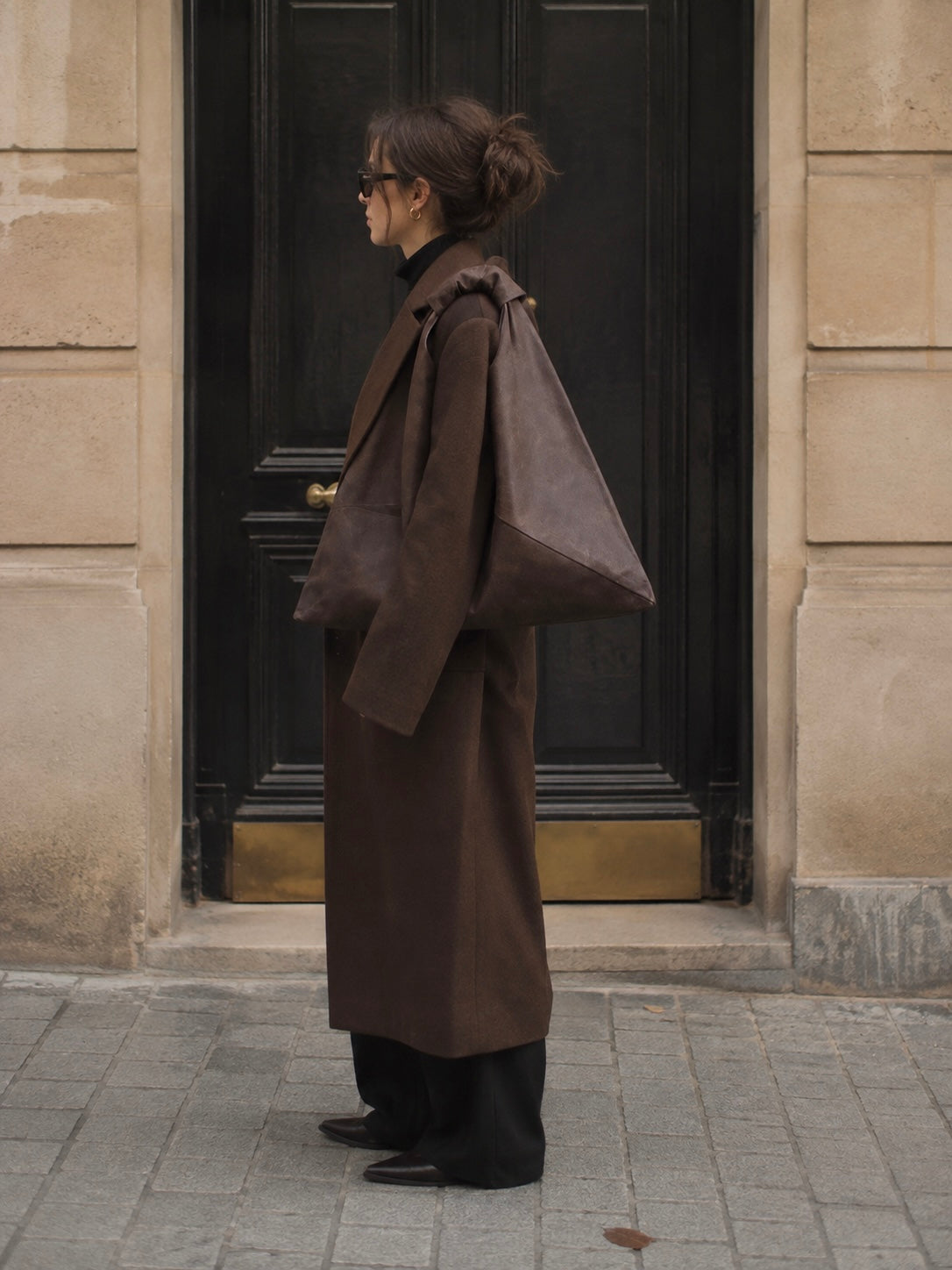 A person stands in profile in front of a dark doorway, wearing a long brown coat and carrying a large brown waxed canvas shoulder bag. Esra Kayira.