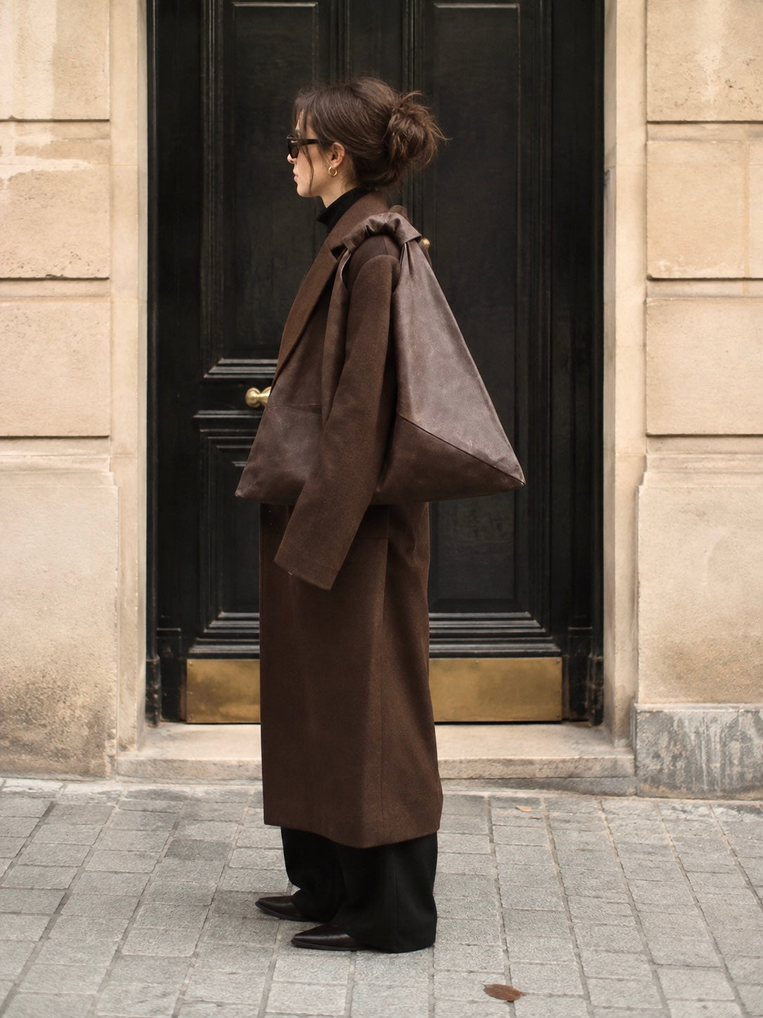 A woman stands in profile in front of a dark wooden door and beige stone building, wearing a long brown coat, black pants, and carrying a large dark brown waxed canvas shoulder bag by Esra Kayira.