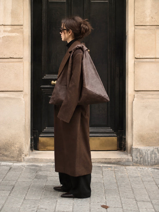 A woman stands in profile in front of a dark wooden door and beige stone building, wearing a long brown coat, black pants, and carrying a large dark brown waxed canvas shoulder bag by Esra Kayira.