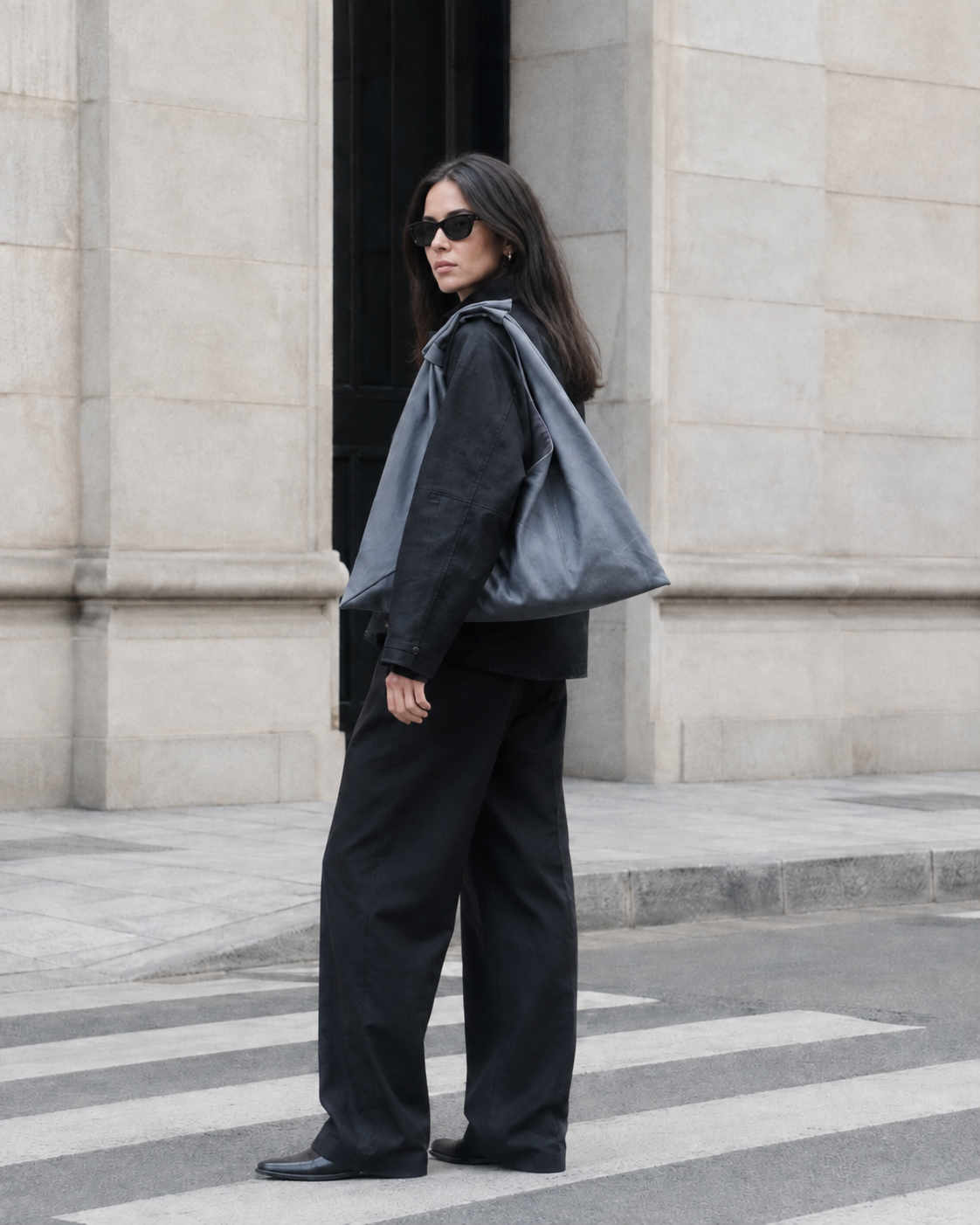 A woman wearing a black outfit, sunglasses, and a large grey waxed canvas origami shoulder bag stands on a city crosswalk in front of a stone building. Bag by Esra Kayira.